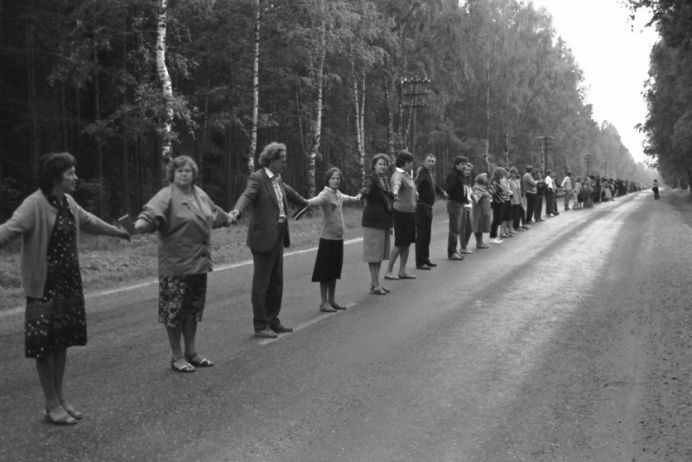 Menschen stehen nebeneinander auf dem Mittelstreifen einer Landstra&szlig;e und halten sich an den H&auml;nden.