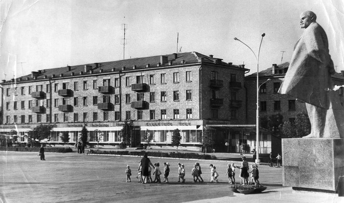 Foto: Öffentlicher, von Gebäuden gesäumter Platz mit Lenin-Denkmal am rechten Rand über den eine Gruppe Kinder mit Betreuerinnen geht.