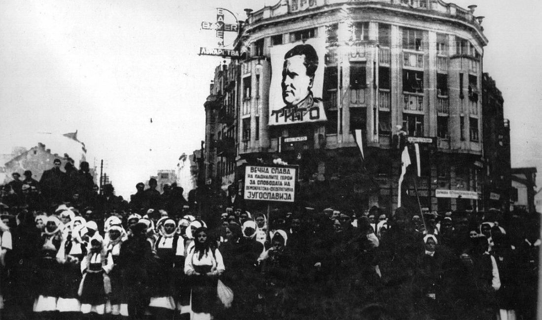 Menschenmenge, teilweise in Tracht, mit Spruchplakaten vor einem Stadthaus mit Tito-Banner.