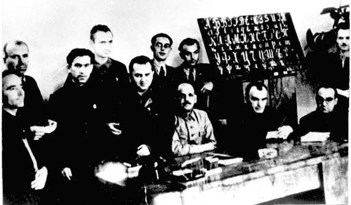 Foto: Gruppe sitzender und stehender Männer im Raum neben einer Buchstabentafel.